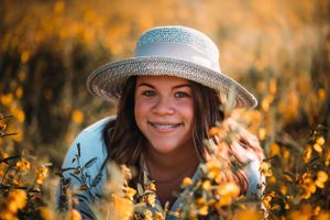 Smiling teenage girl with braces wearing a sun hat lying in a golden yellow flower field.