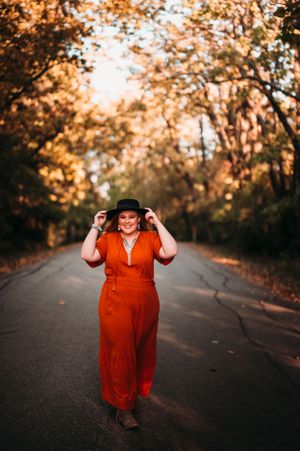 Woman in orange dress and black hat walking down tree-lined road in autumn foliage.