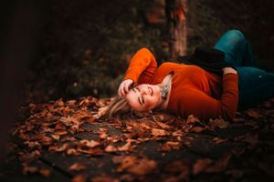 Young woman in orange sweater lying in fallen autumn leaves on ground.