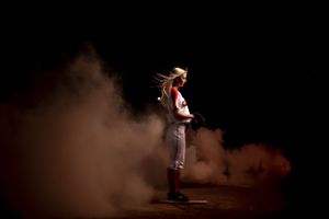 Female softball pitcher in uniform stands dramatically surrounded by smoke on a dark dramatic sports portrait.
