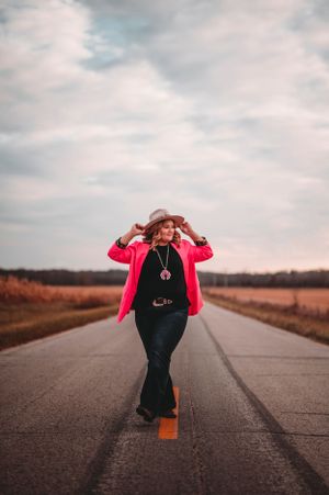 Woman in pink blazer and hat walking down rural road at sunset.