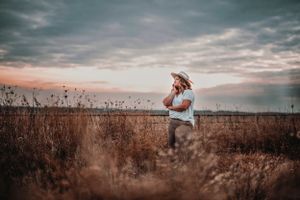 Woman in straw hat standing in golden field at sunset holding phone.