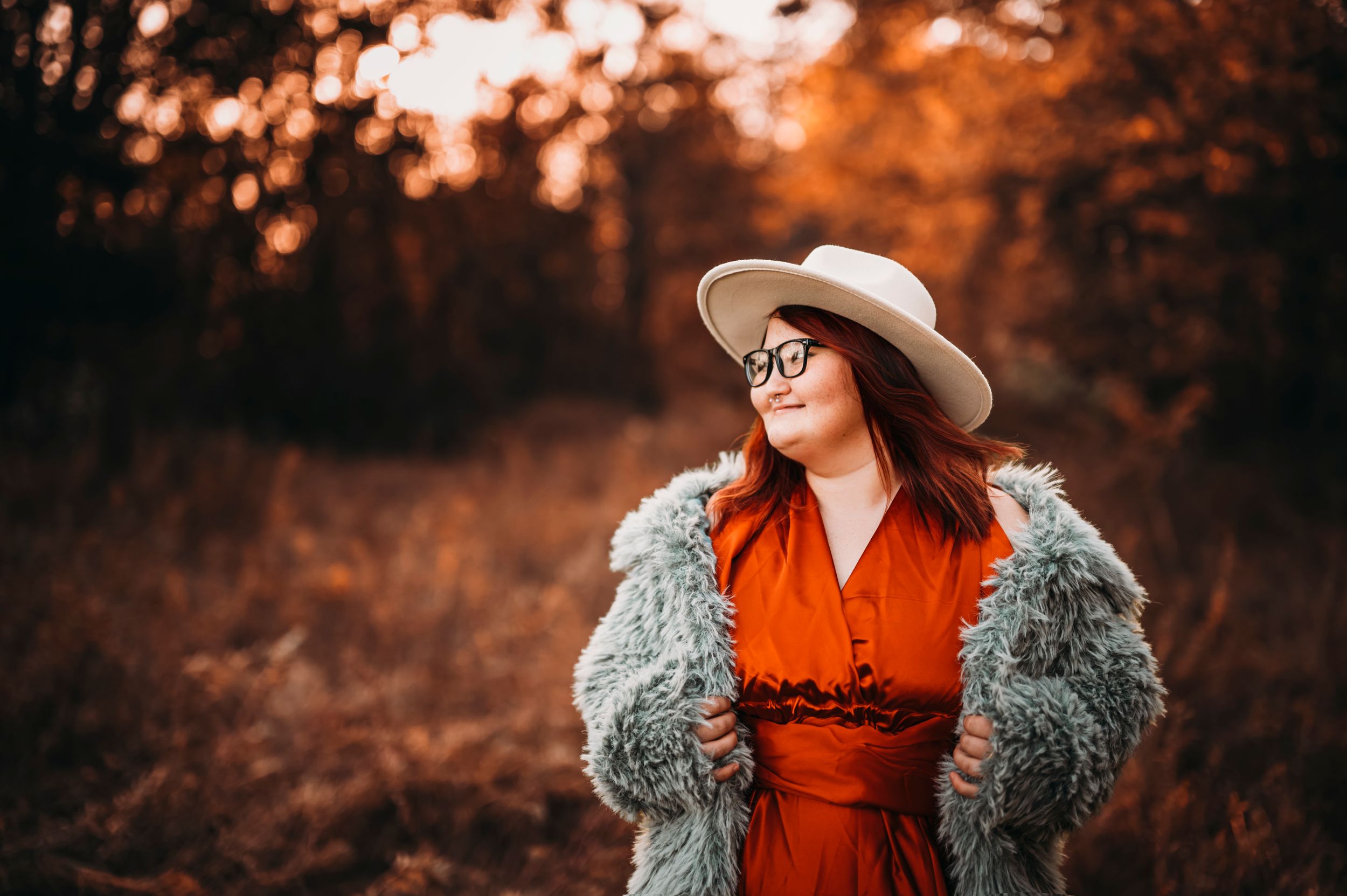 Woman in orange dress fur coat and white hat posing in autumn forest.