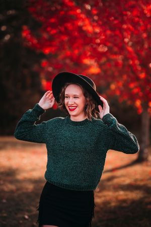 Smiling woman in green sweater and black hat posing before vibrant red autumn foliage.