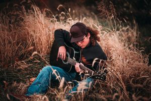Musician playing acoustic guitar while sitting in tall golden grass field.