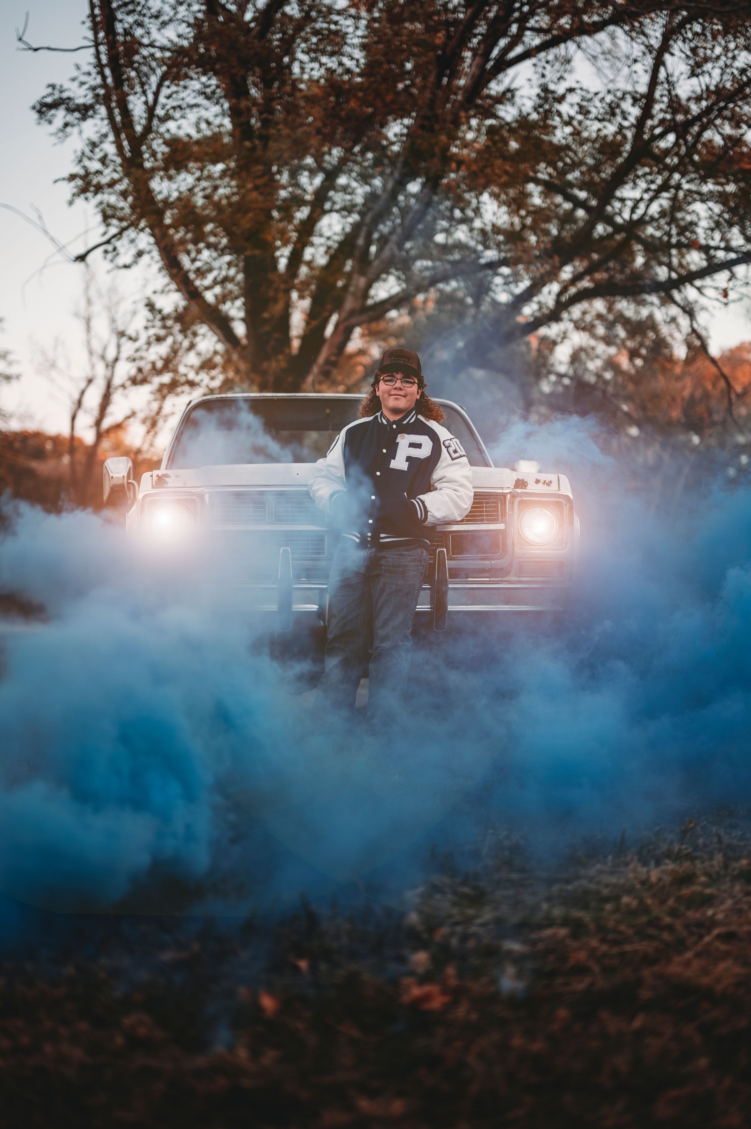 Person in varsity jacket leaning on truck hood surrounded by blue smoke at golden hour.