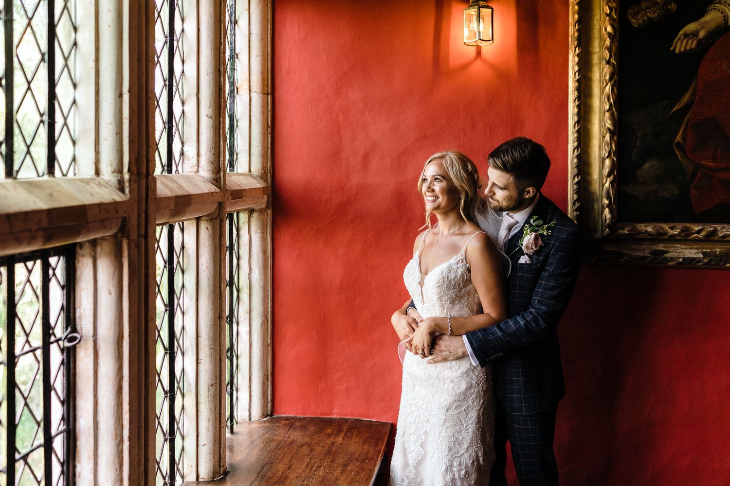 Couple in wedding attire stand by ornate window with red walls.