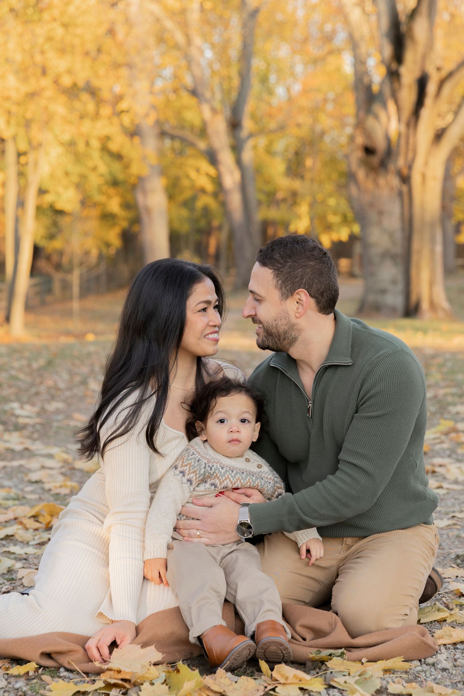 family of three sitting on the ground smiling at each other in front of tree trees in autumn