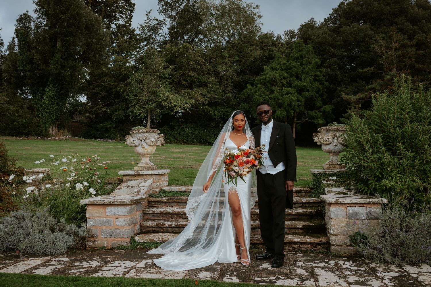 Wedding couple poses on stone steps with lush garden backdrop and flowing cathedral veil.