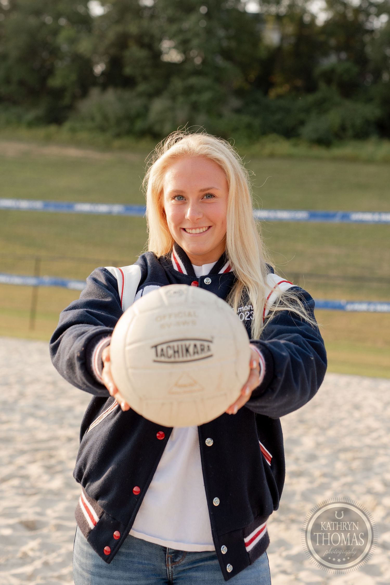 Student athlete holds volleyball while wearing navy varsity jacket on sandy court.
