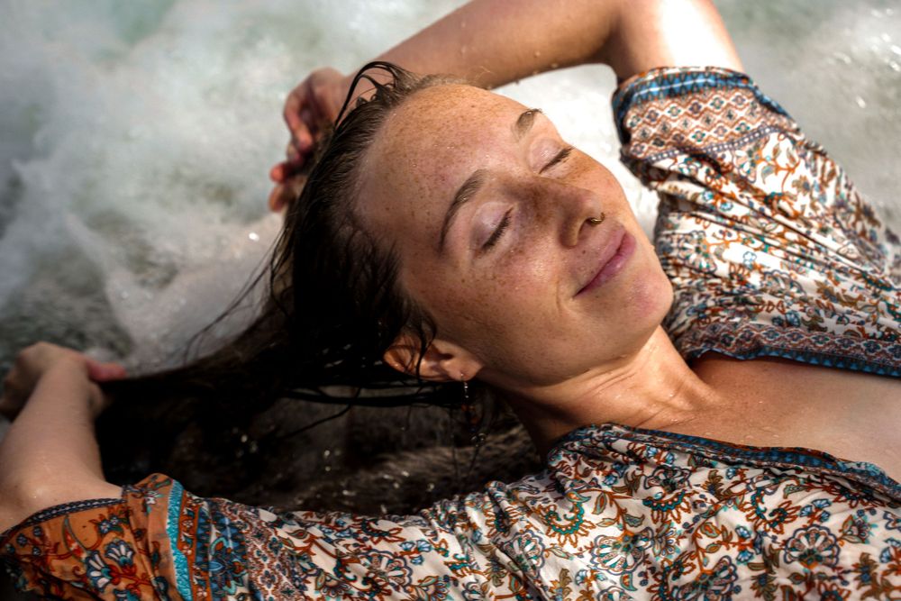 Person lying by water wearing patterned dress with peaceful expression in sunlight.