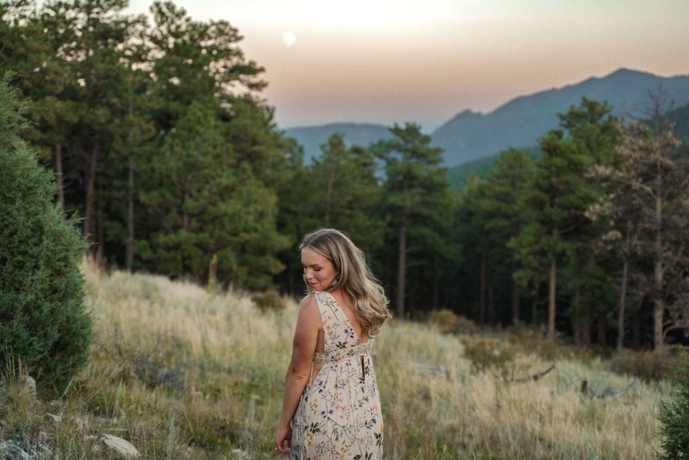 Floral dress-clad figure stands in golden mountain meadow at sunset with pine trees in background.