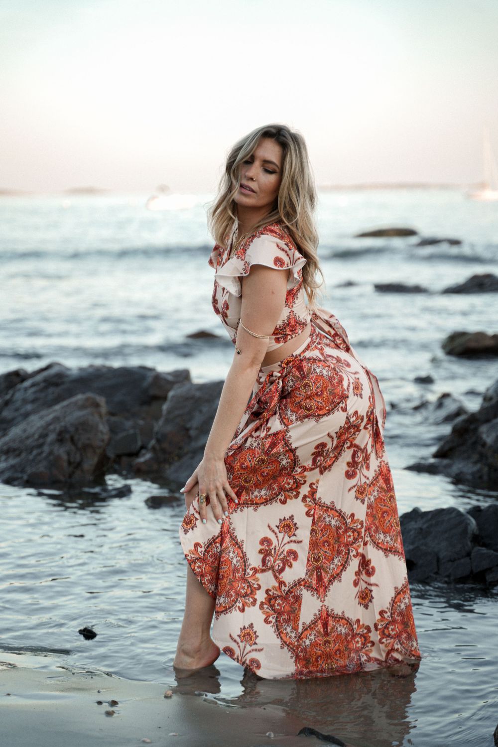 Woman in red and white floral dress plays in ocean waves near rocky shore.