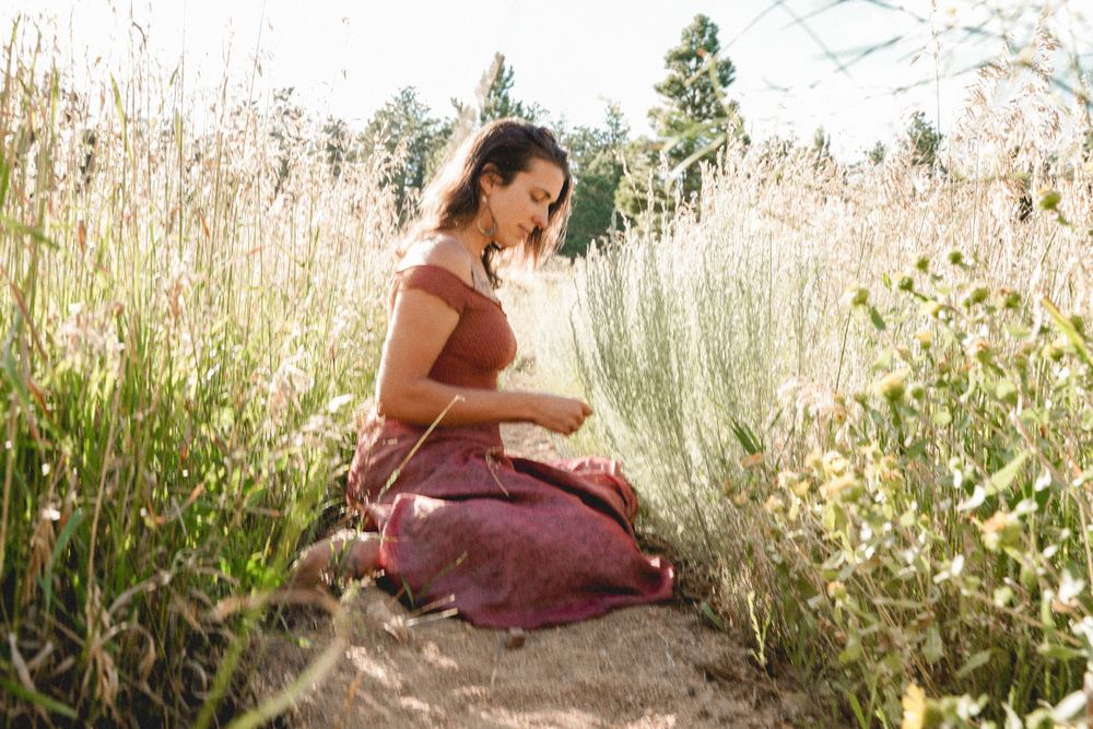 Person in burgundy dress sits peacefully among sunlit wildflowers and tall grasses.