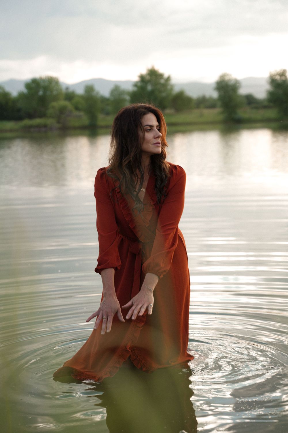 A person in a flowing red dress stands in tranquil lake water at sunset with mountains in the background.