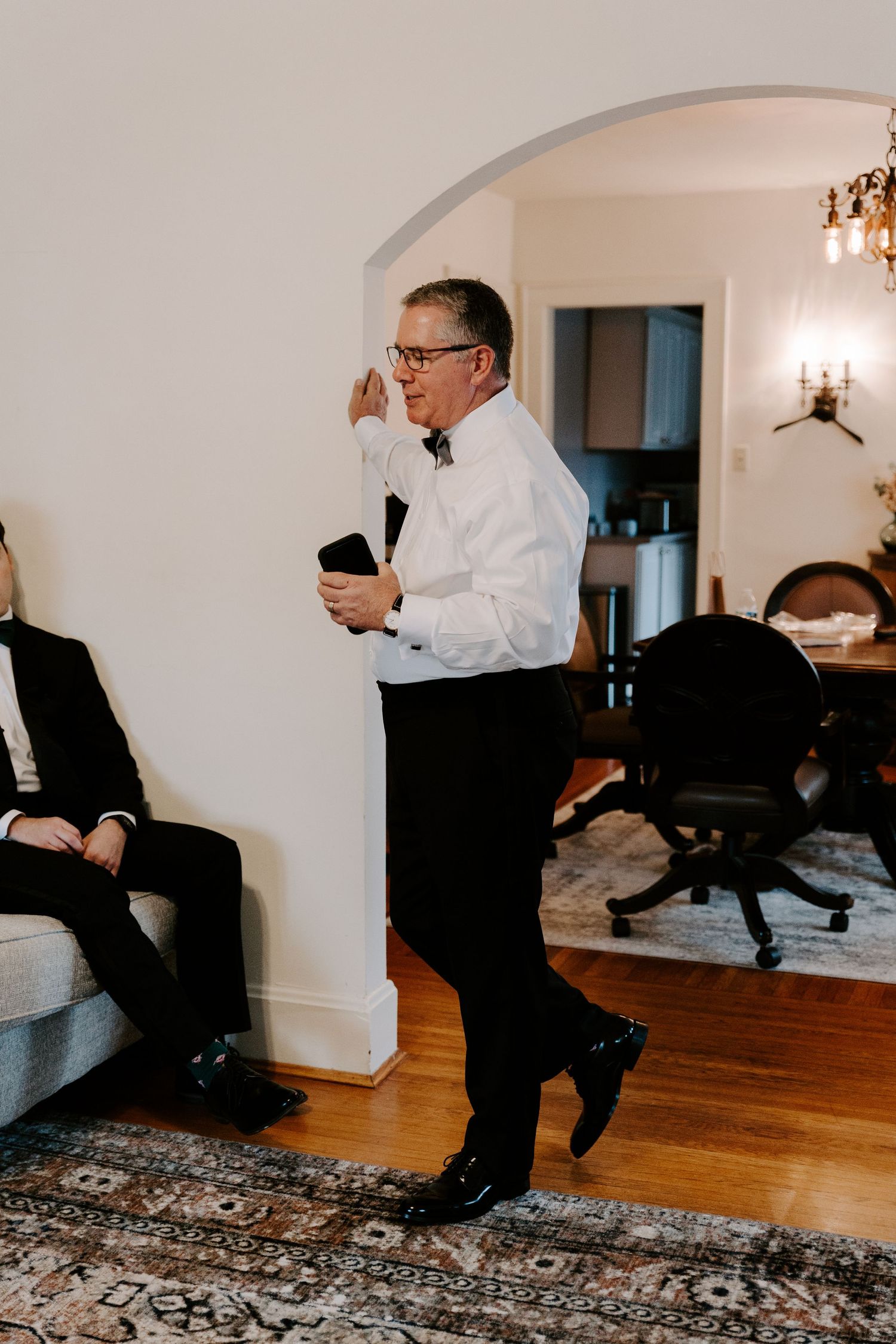 Man in formal attire speaking while standing on hardwood floors in a bright room.