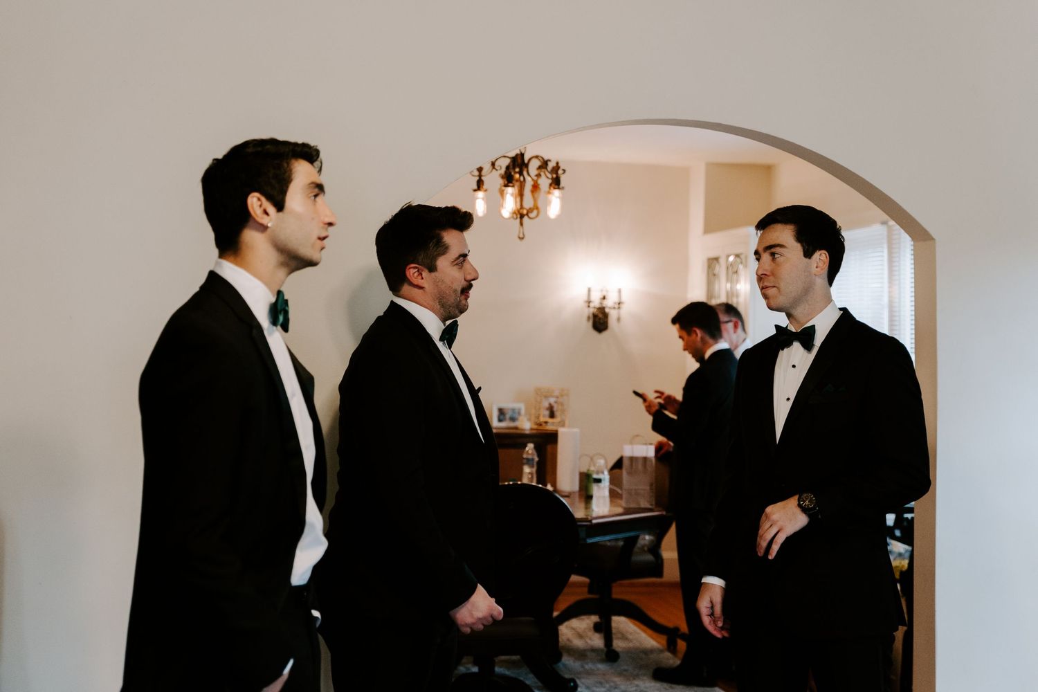 Groomsmen in black tuxedos gathered in conversation under an arched doorway.
