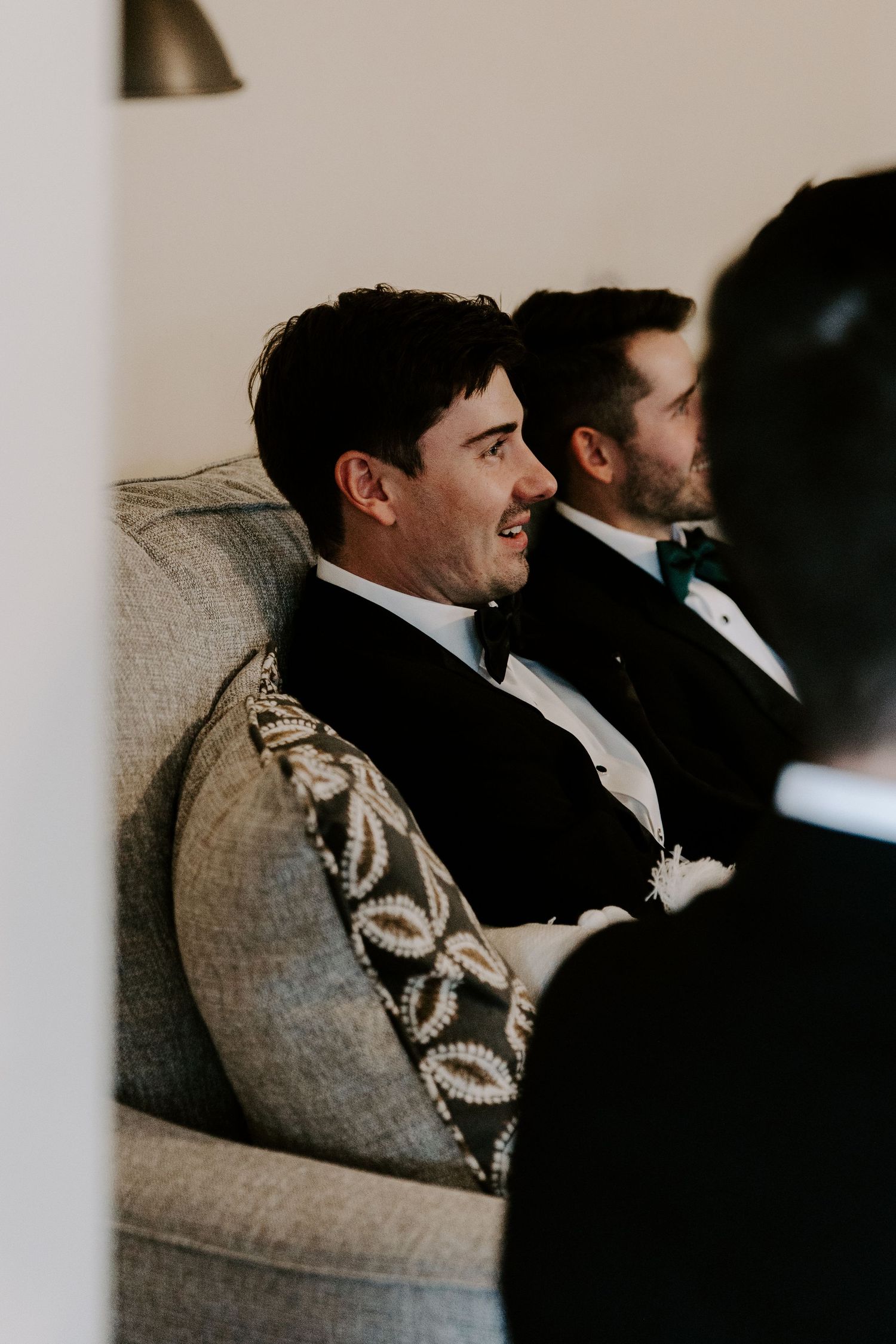 Wedding party member in tuxedo smiling while seated on a gray couch.