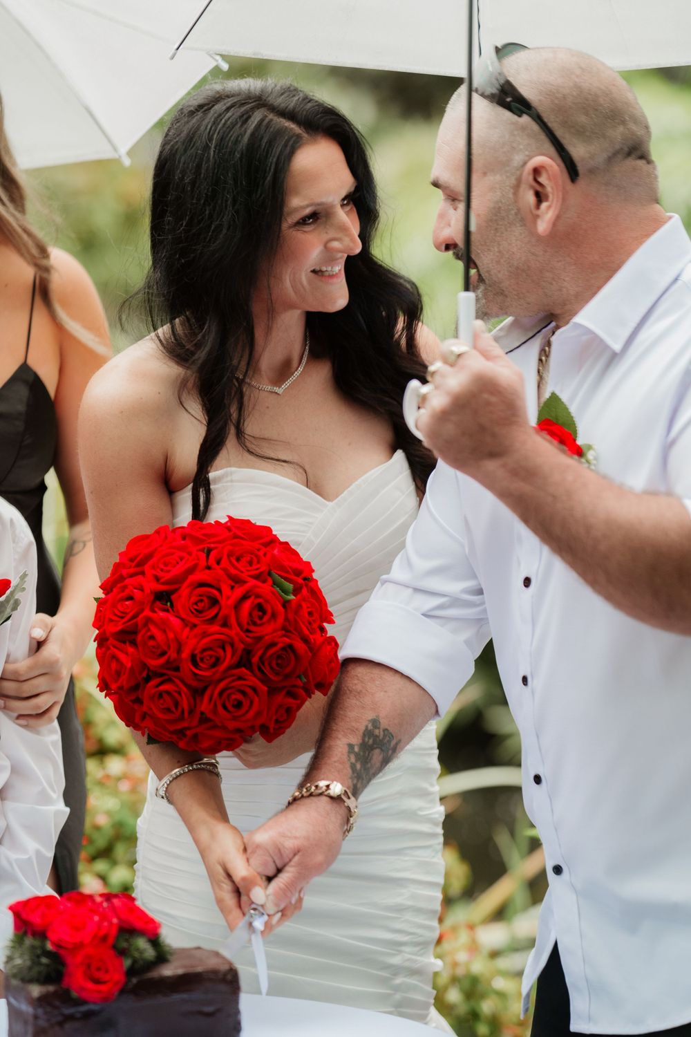 A couple embraces during a wedding photoshoot with the bride holding a vibrant red rose bouquet.