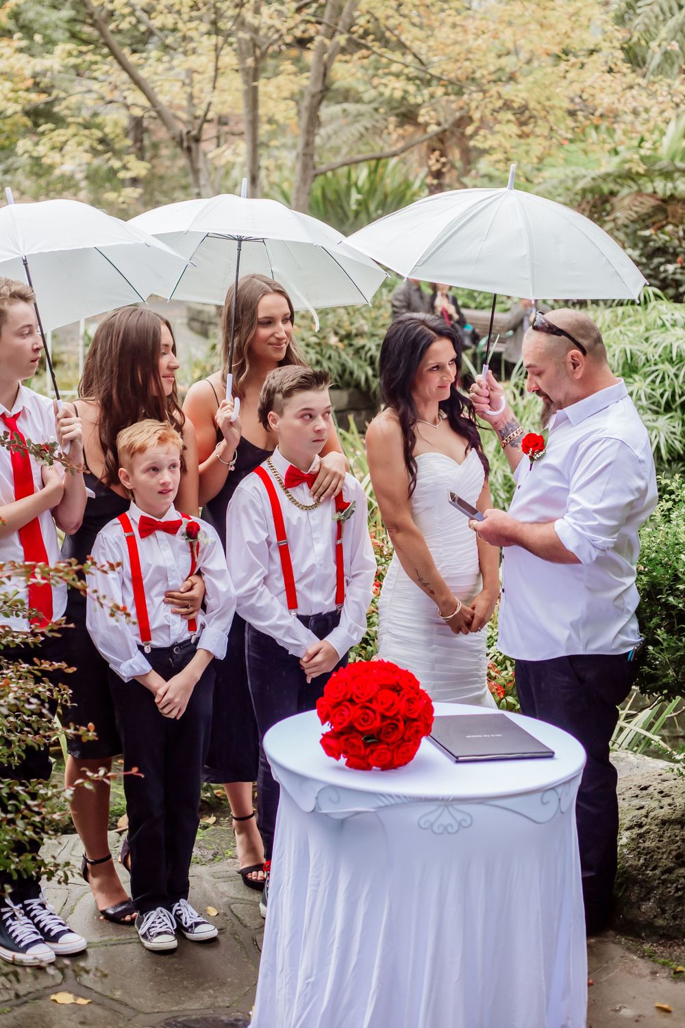Wedding party with children in red bowties poses together under white umbrellas.