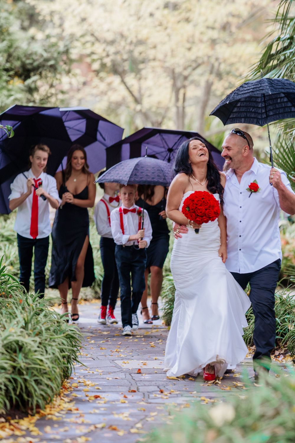 Wedding party walks under purple umbrellas on a rainy day with red accents.