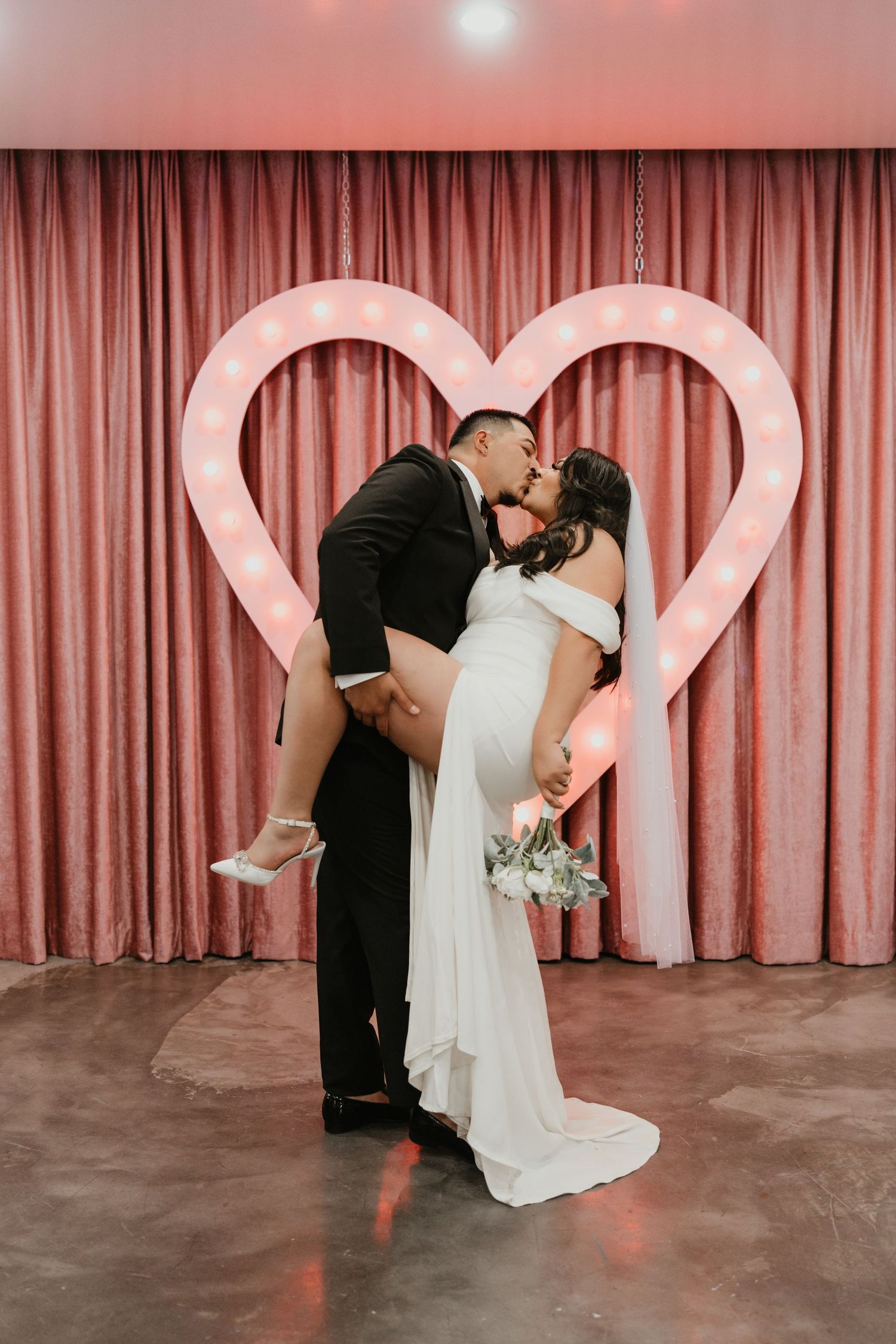 Romantic wedding photo of couple kissing in front of pink heart backdrop during reception.