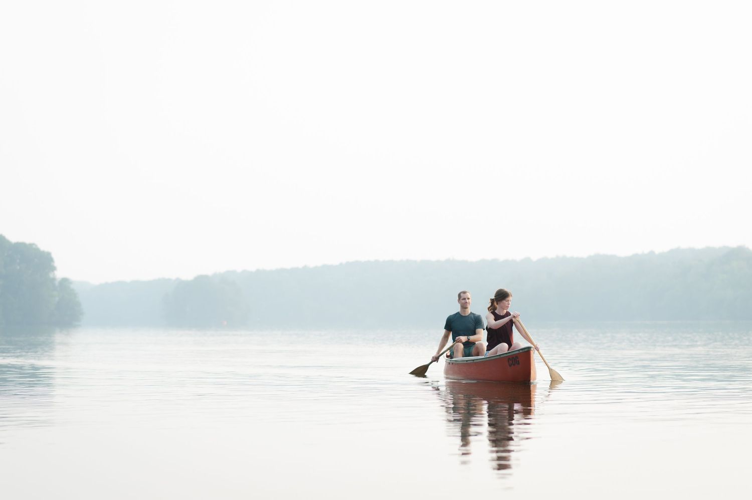 Two people paddling in a canoe on a misty lake during early morning.