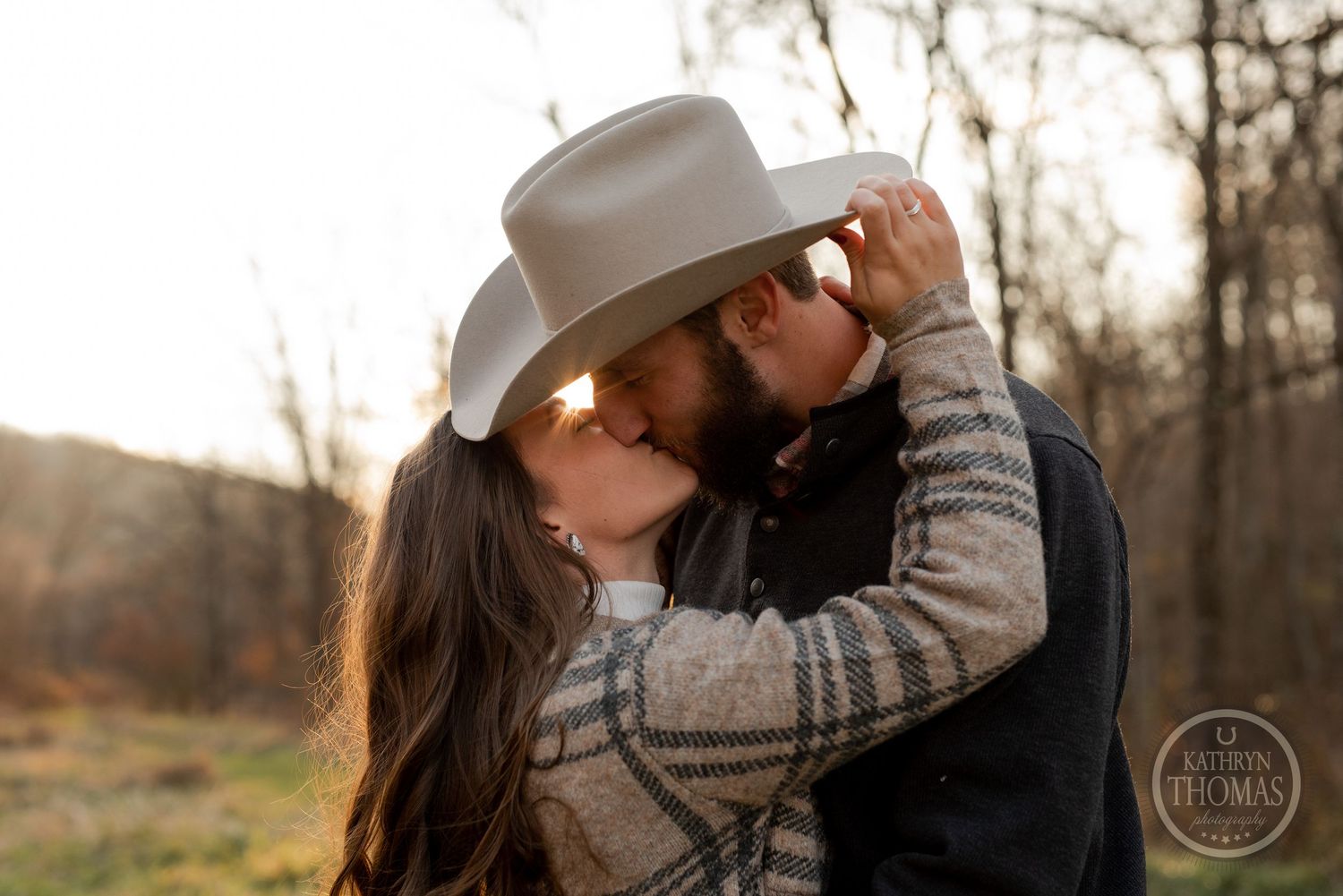 A couple embraces in an autumn field during golden hour while sharing a cowboy hat.
