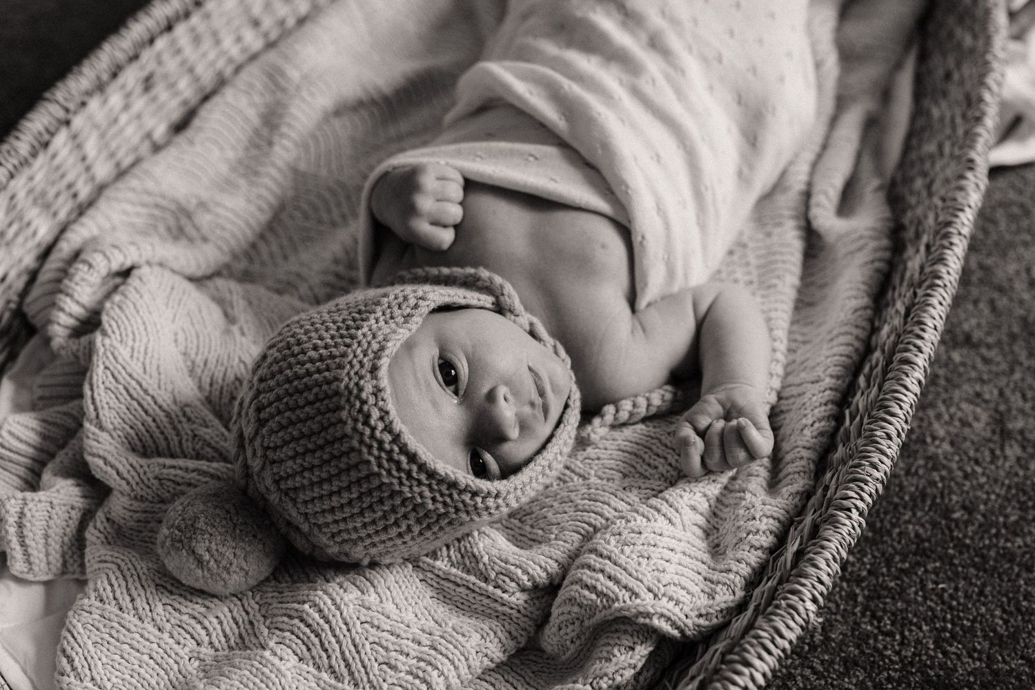 Newborn baby wearing knit bonnet lies peacefully on white blanket in black and white portrait.
