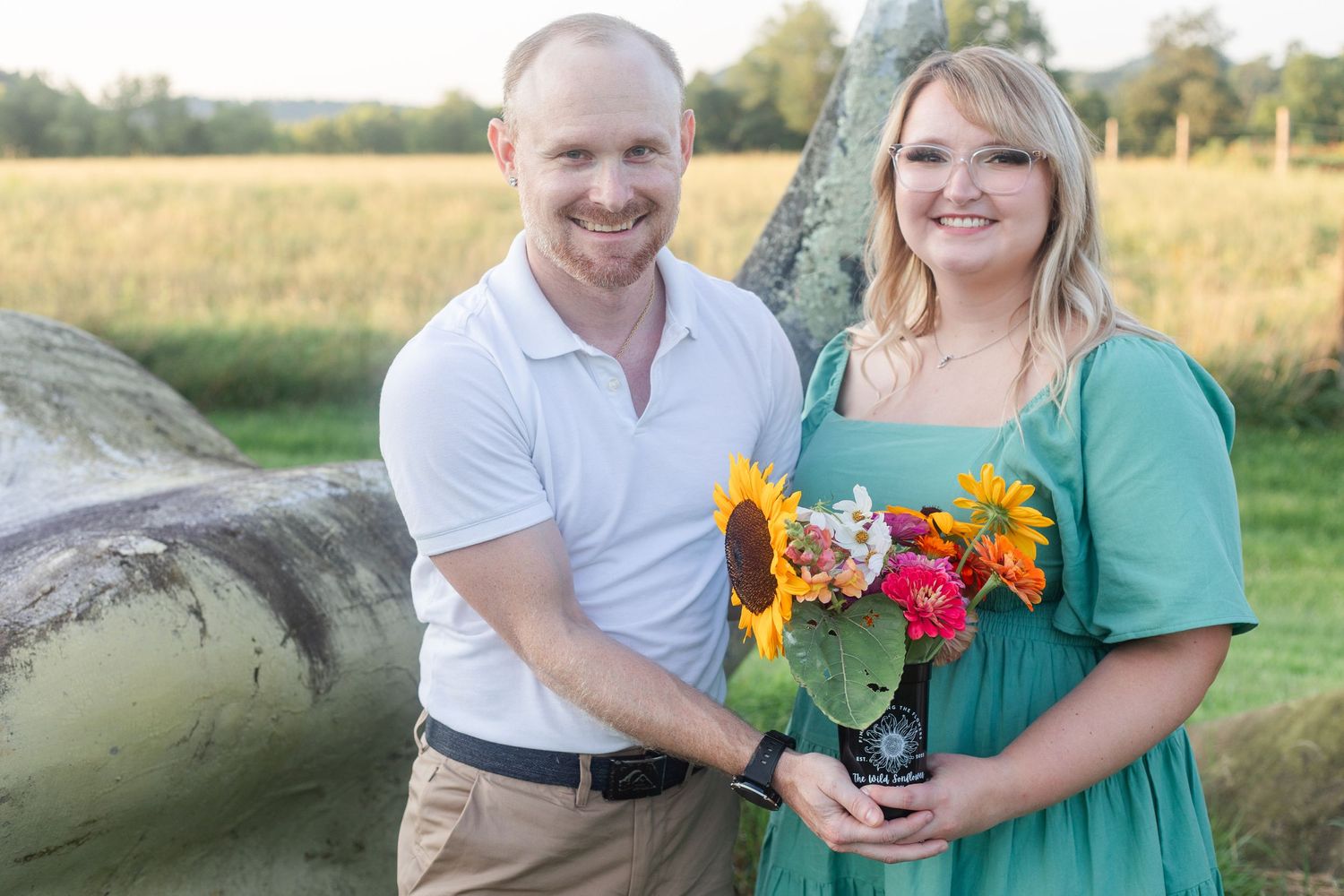 A couple poses outdoors with bright sunflowers in a green field setting.