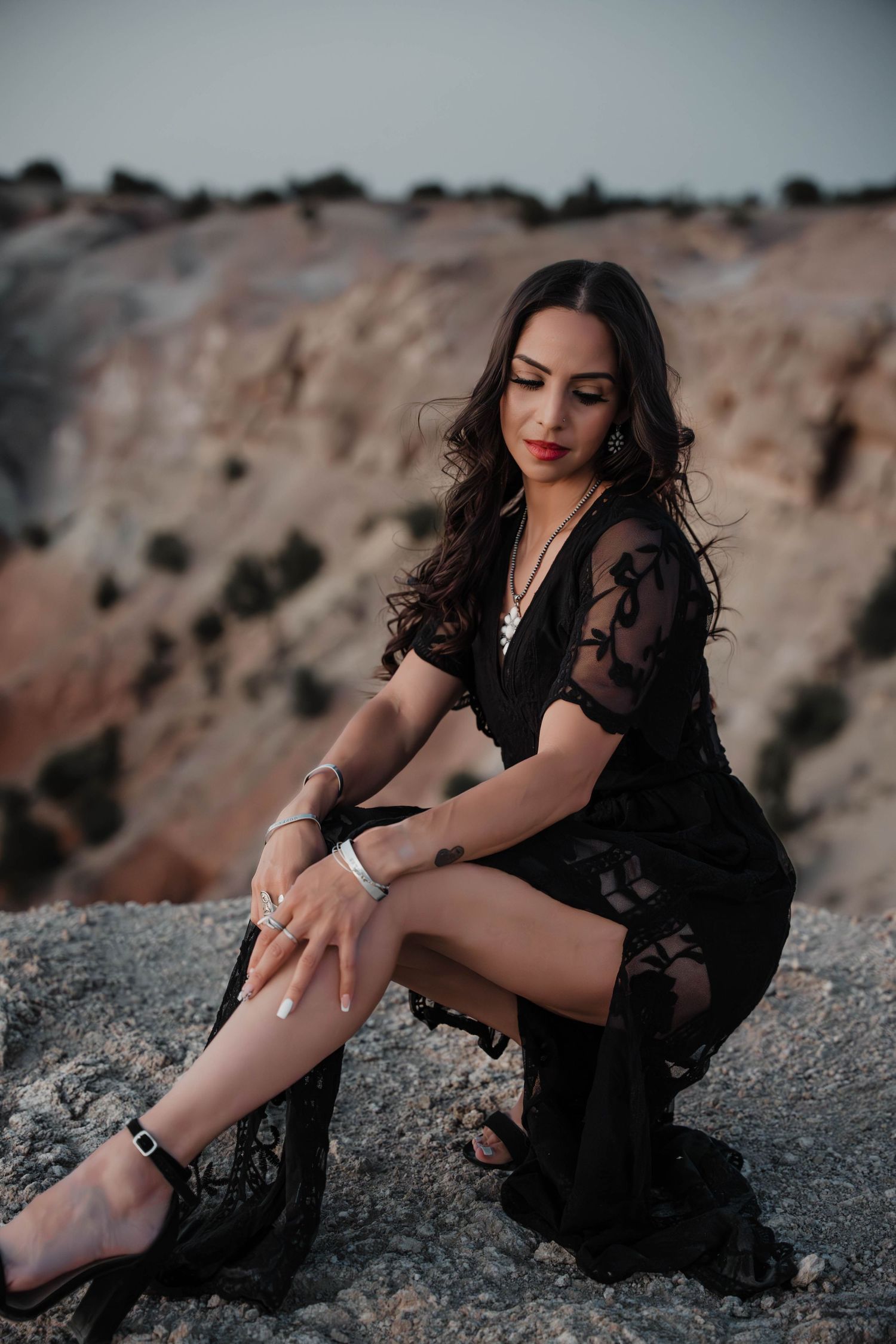 Woman in black lace dress poses on rocky terrain with desert backdrop.