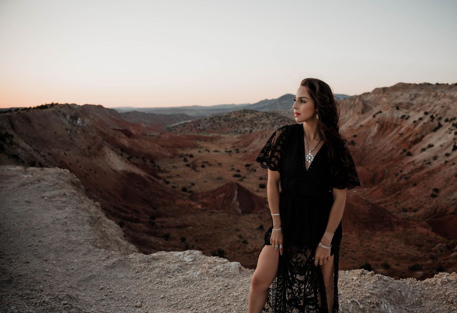 Woman in black dress stands on cliff edge overlooking red rock canyon vista.