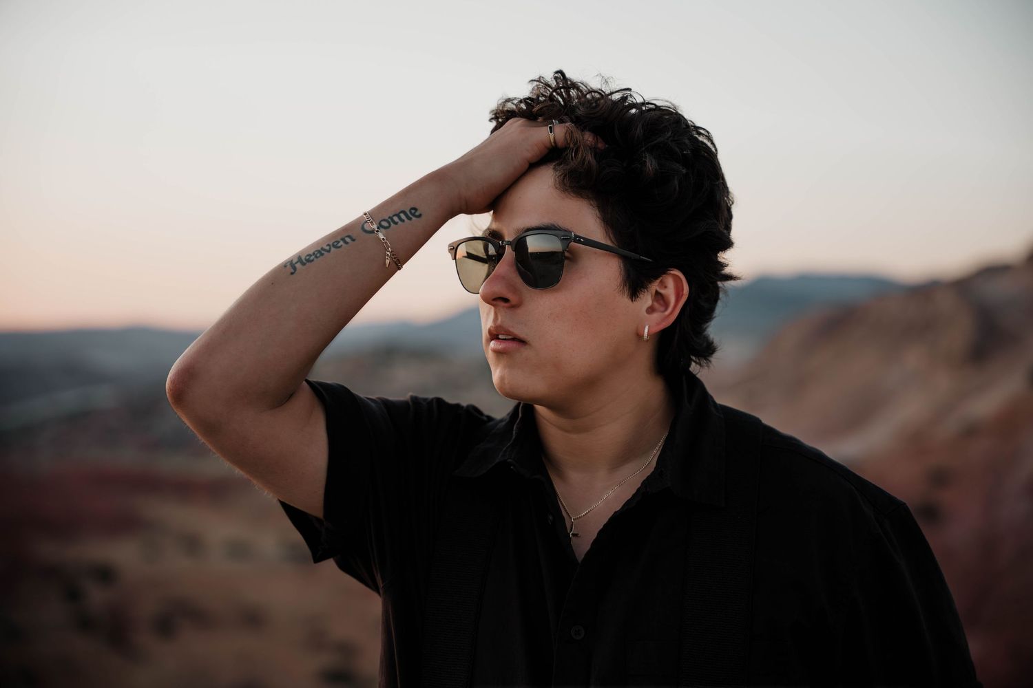 Figure in black attire shields eyes while posing against rocky desert terrain.