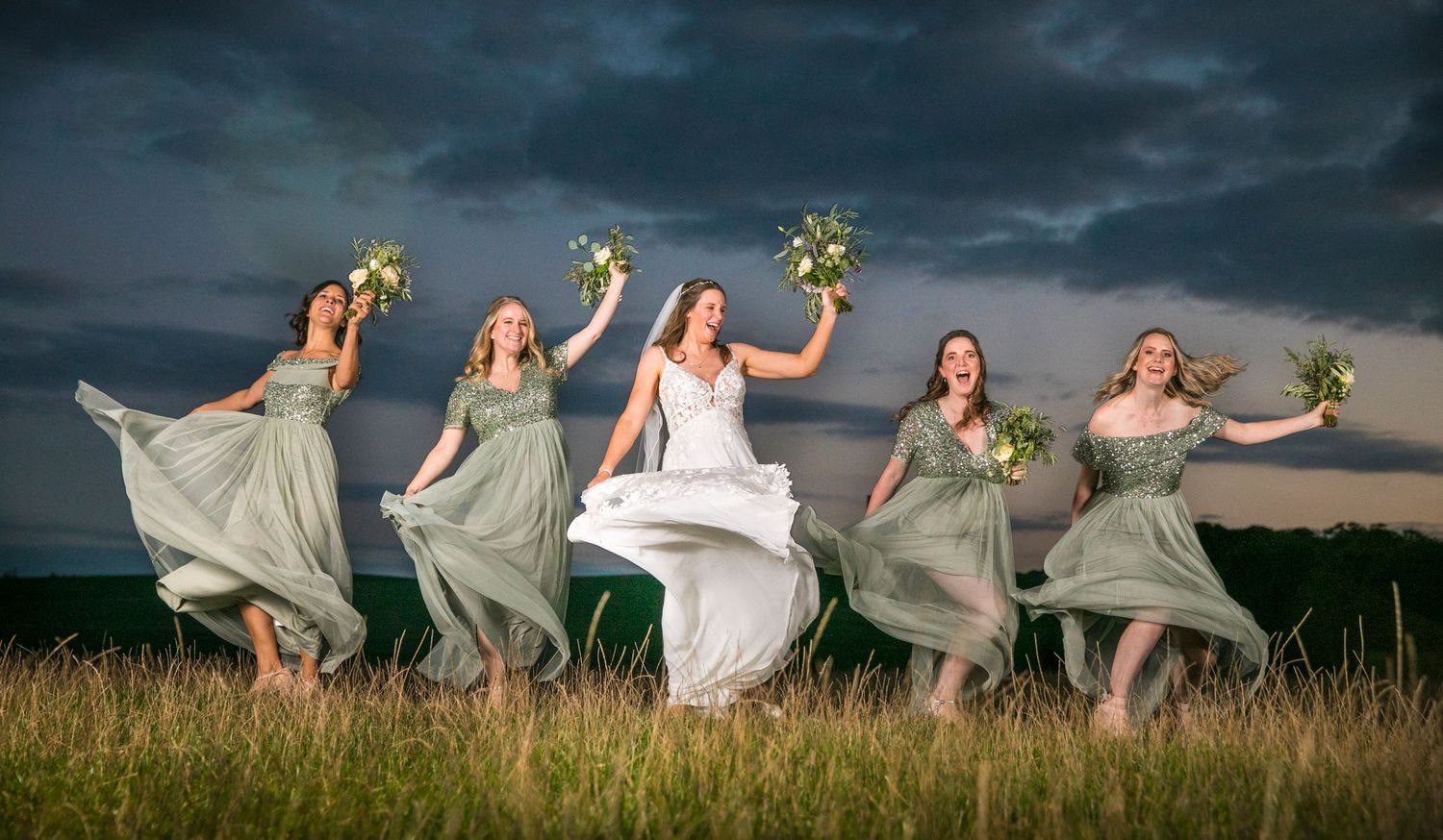 Bridal party dances joyfully in a field at dusk with dresses flowing in the wind.