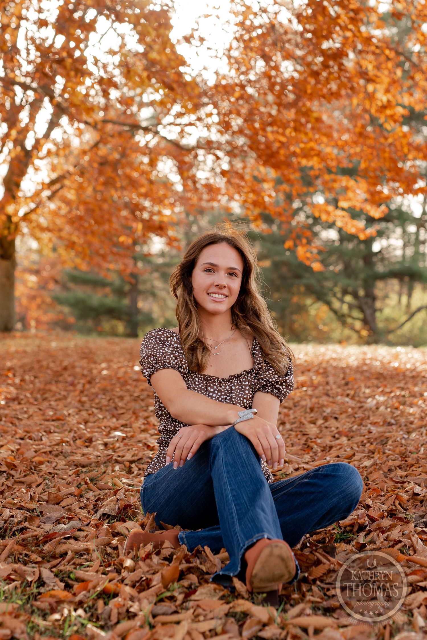 A relaxed portrait sitting cross-legged on fallen autumn leaves wearing jeans and a patterned top.