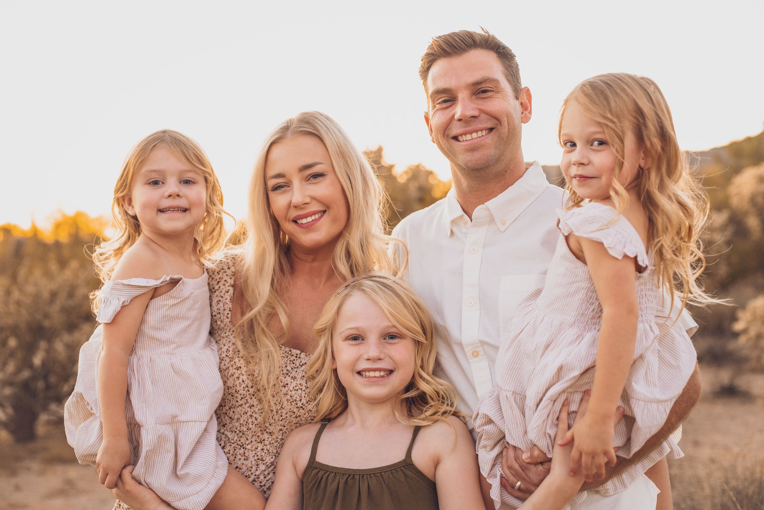 A family portrait taken outdoors against a light background wearing coordinated neutral colors.
