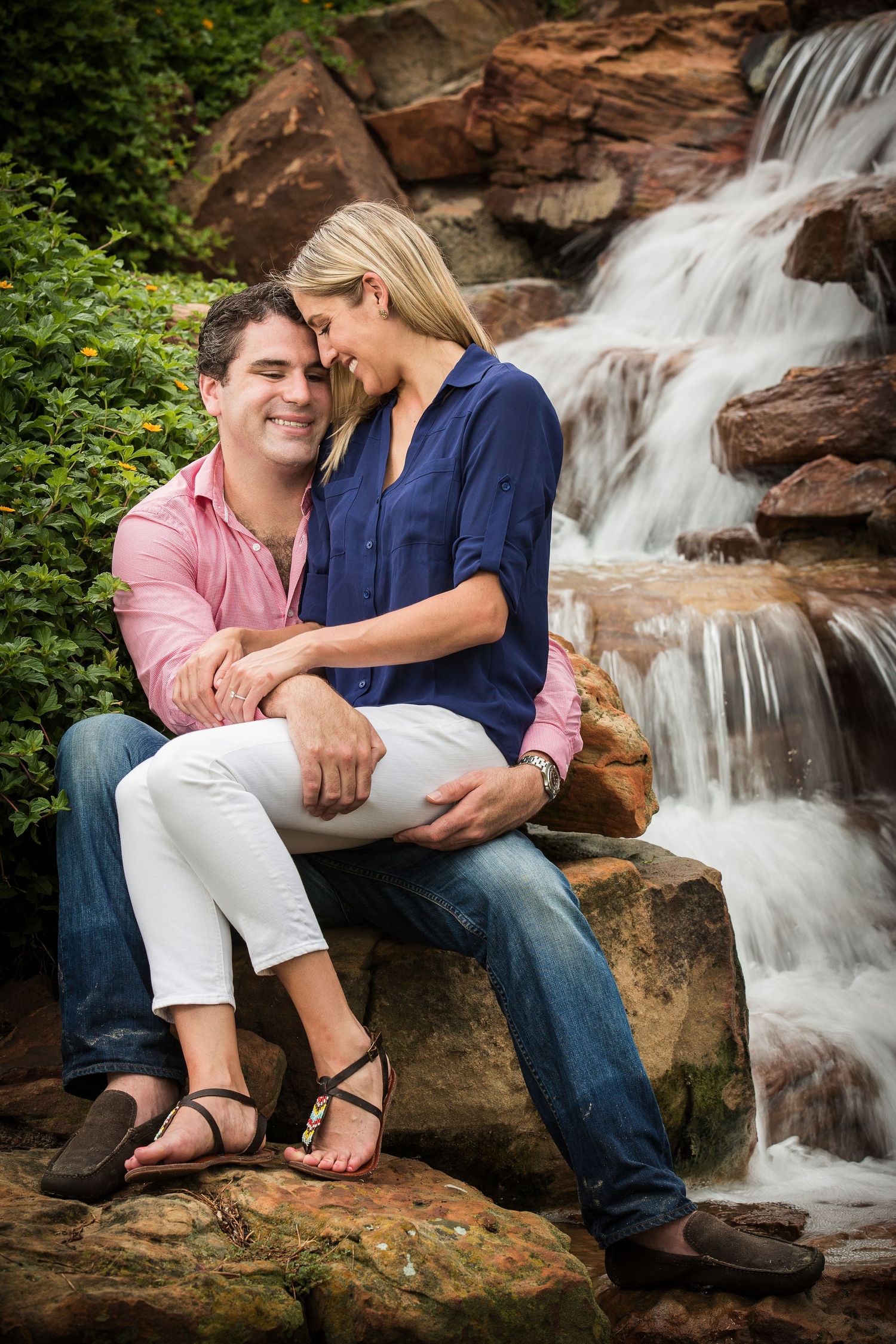 A couple sits intimately on rocks beside a cascading waterfall in a romantic outdoor setting.