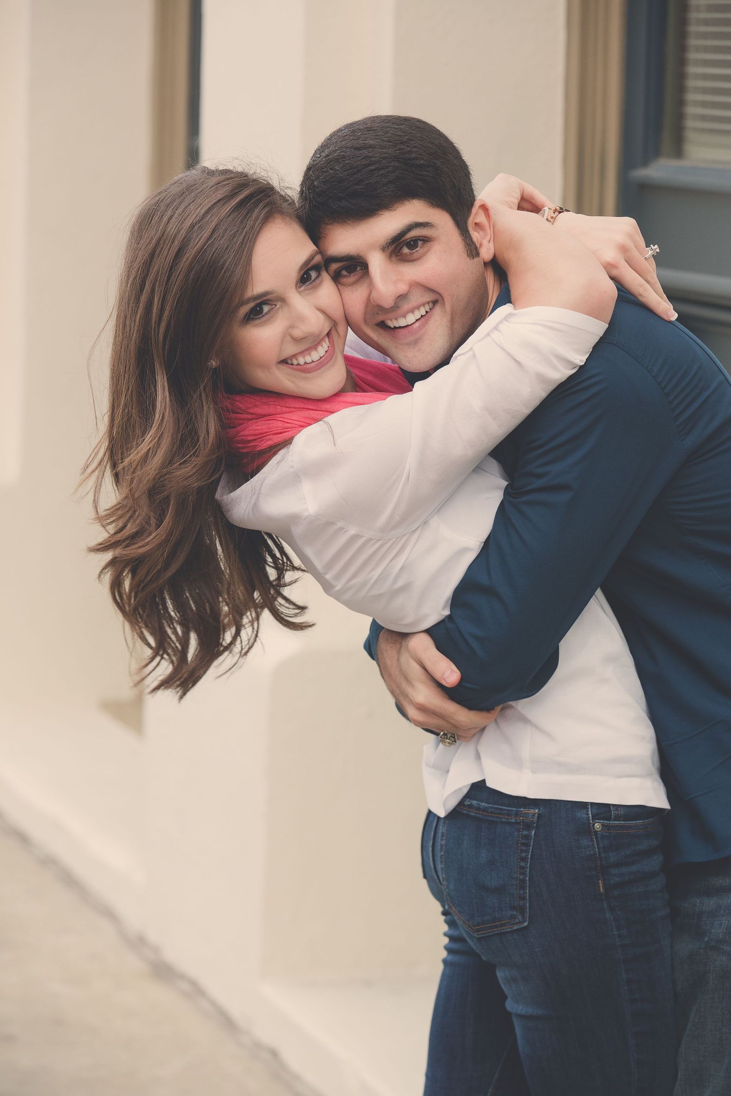 A playful couple embraces against a light colored wall wearing casual attire.