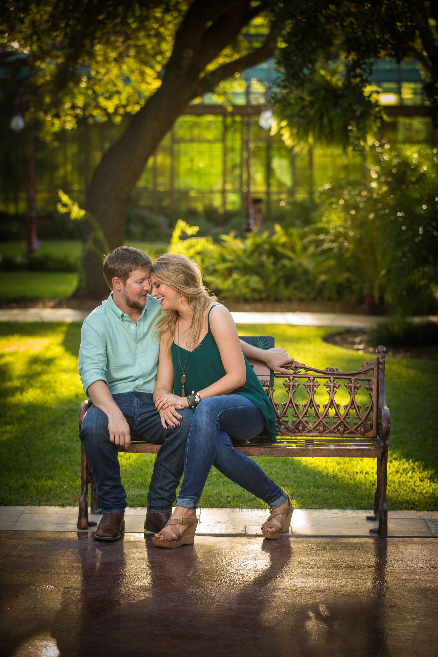 Couple sitting close together on a wooden park bench surrounded by lush greenery in warm sunlight.