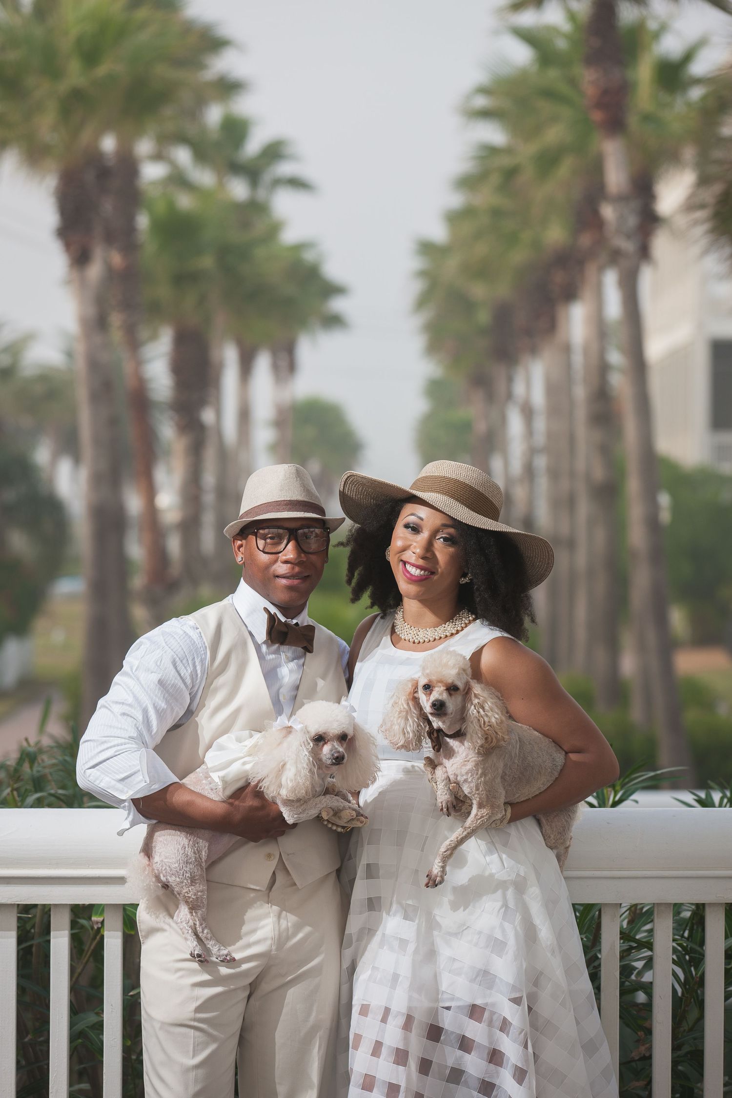Elegant couple in white attire posing with two small white dogs on a palm tree-lined balcony.