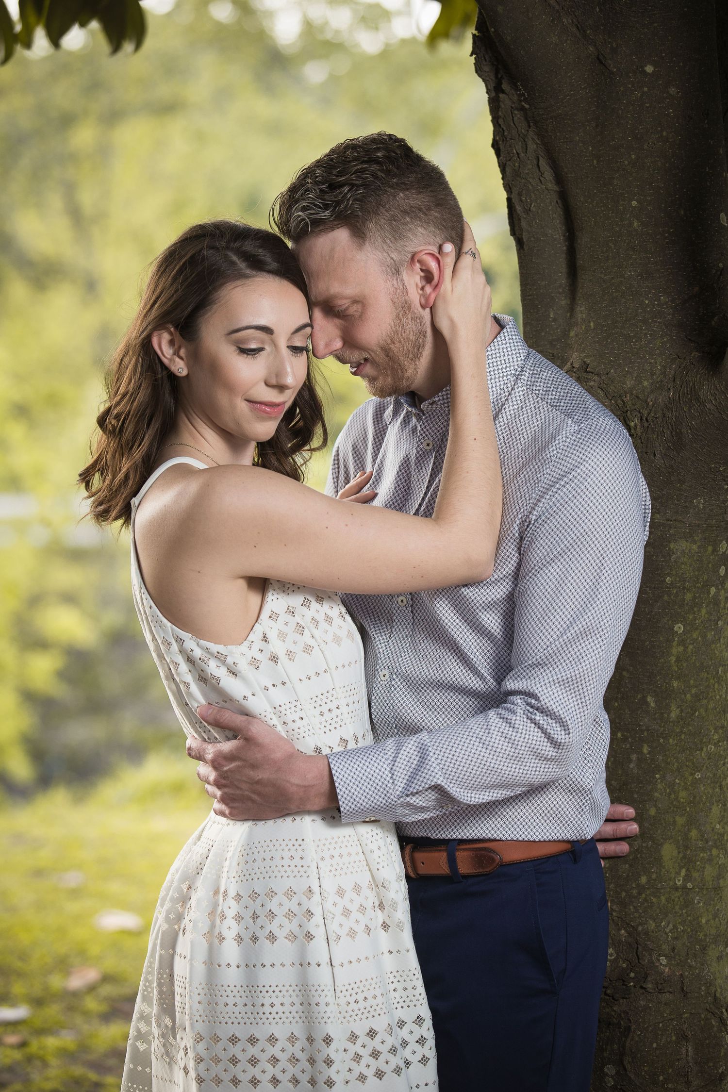Couple in casual attire sharing an intimate embrace under a tree in soft lighting.
