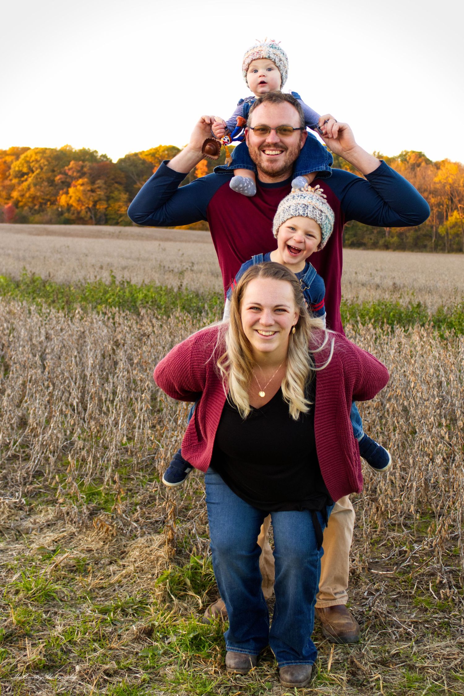 Family of four posing next a soybean field. Warm sunrays are cast over the fall foliage behind them. All are smiling.