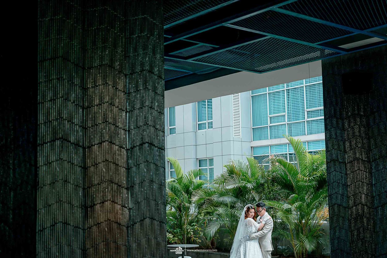 Wedding couple embracing under modern architectural pillars with greenery in background.