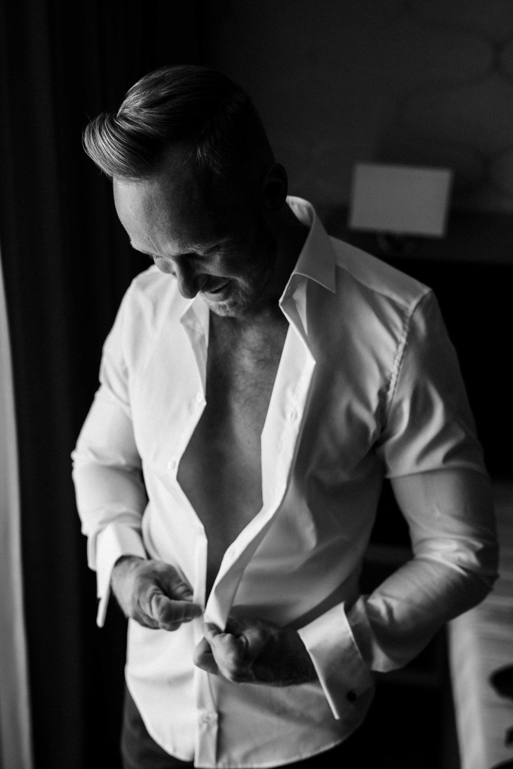 Black and white portrait of groom buttoning crisp white dress shirt.