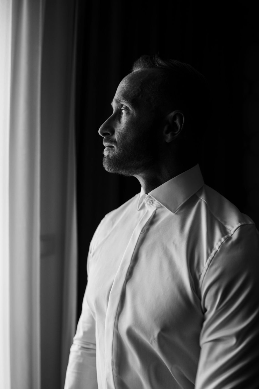 Profile portrait of groom in white shirt gazing thoughtfully by window.
