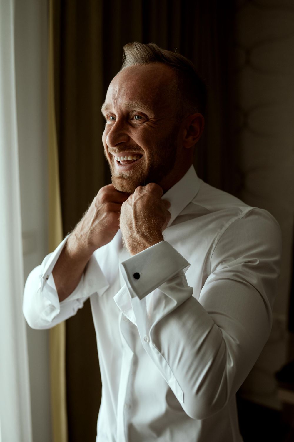 Groom adjusting collar of white dress shirt while smiling near window.