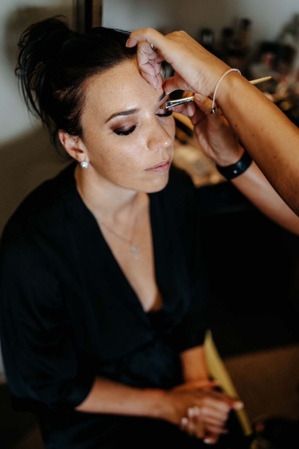 Makeup artist carefully applying eye makeup on client wearing black robe during wedding preparation.