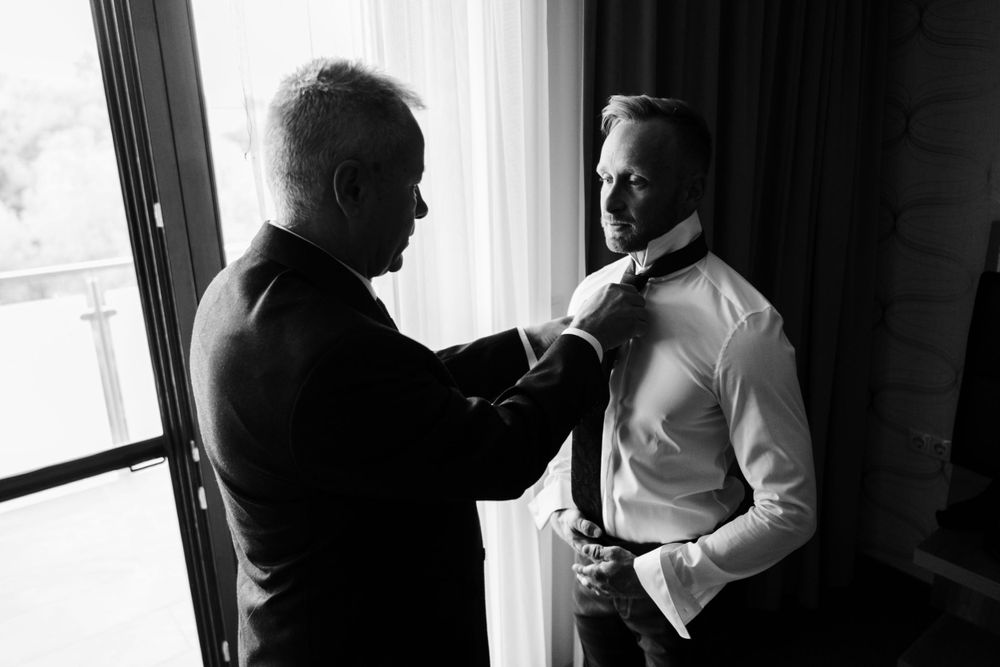 Black and white image of two men adjusting formal attire by window.