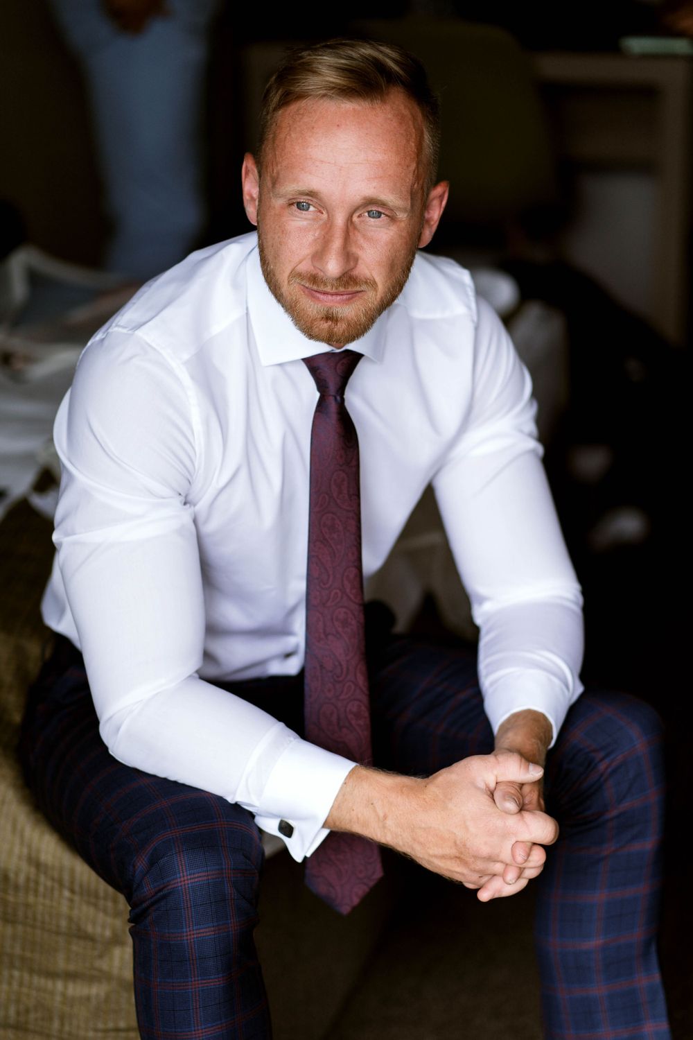 Man in formal business attire sits on edge of bed in plaid dress pants.