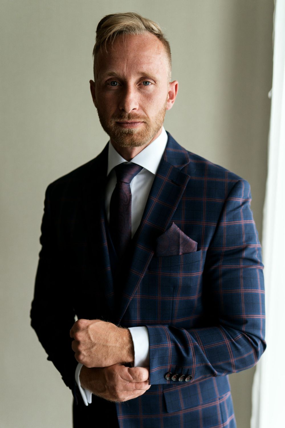 Man poses in navy plaid suit jacket with purple tie against light wall.